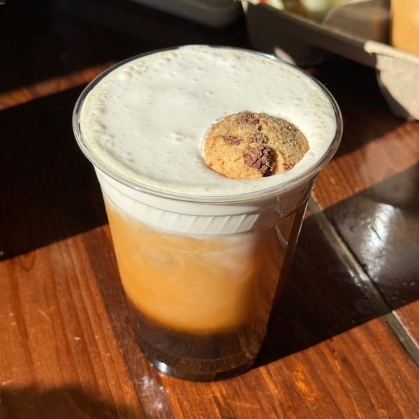 A clear plastic cup of iced coffee with a thick layer of foam and a small cookie on top.