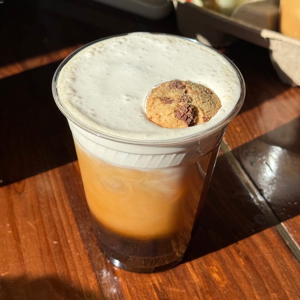 A clear plastic cup of iced coffee with a thick layer of foam and a small cookie on top.