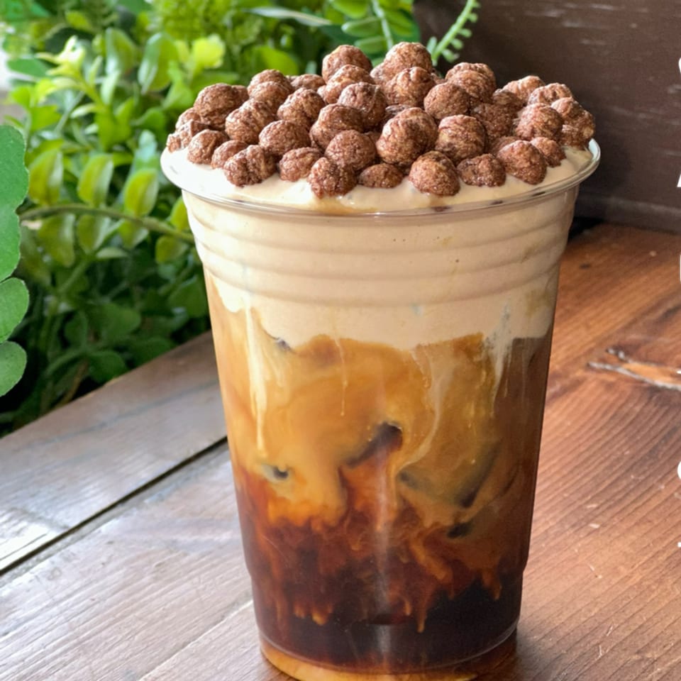 Iced coffee with a creamy top layer and chocolate cereal puffs, on a wooden surface.
