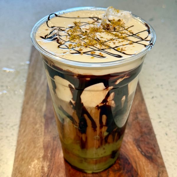 A layered coffee drink with chocolate drizzle, whipped cream, and pistachio crumbles in a clear cup.