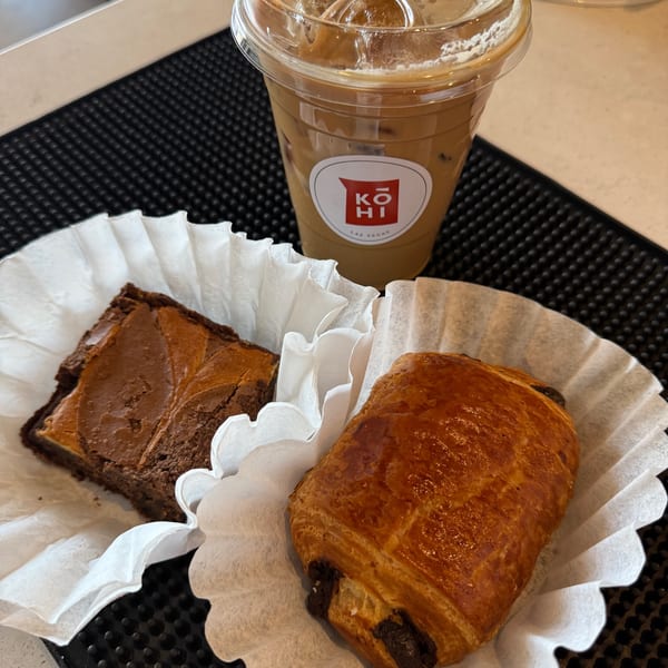 Iced coffee with a Kohi logo, a brownie, and a chocolate croissant on a black mat.