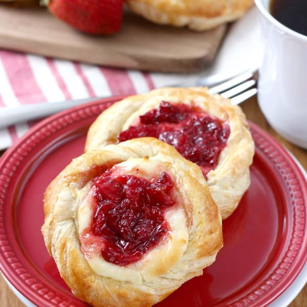 Two cranberry cream cheese danishes on a red plate with coffee and strawberries in the background.
