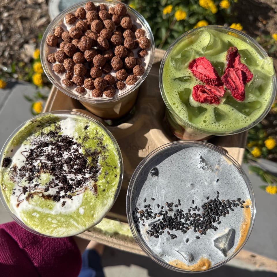 Four colorful iced drinks with various toppings, including cereal, dried strawberries, and black sesame seeds.