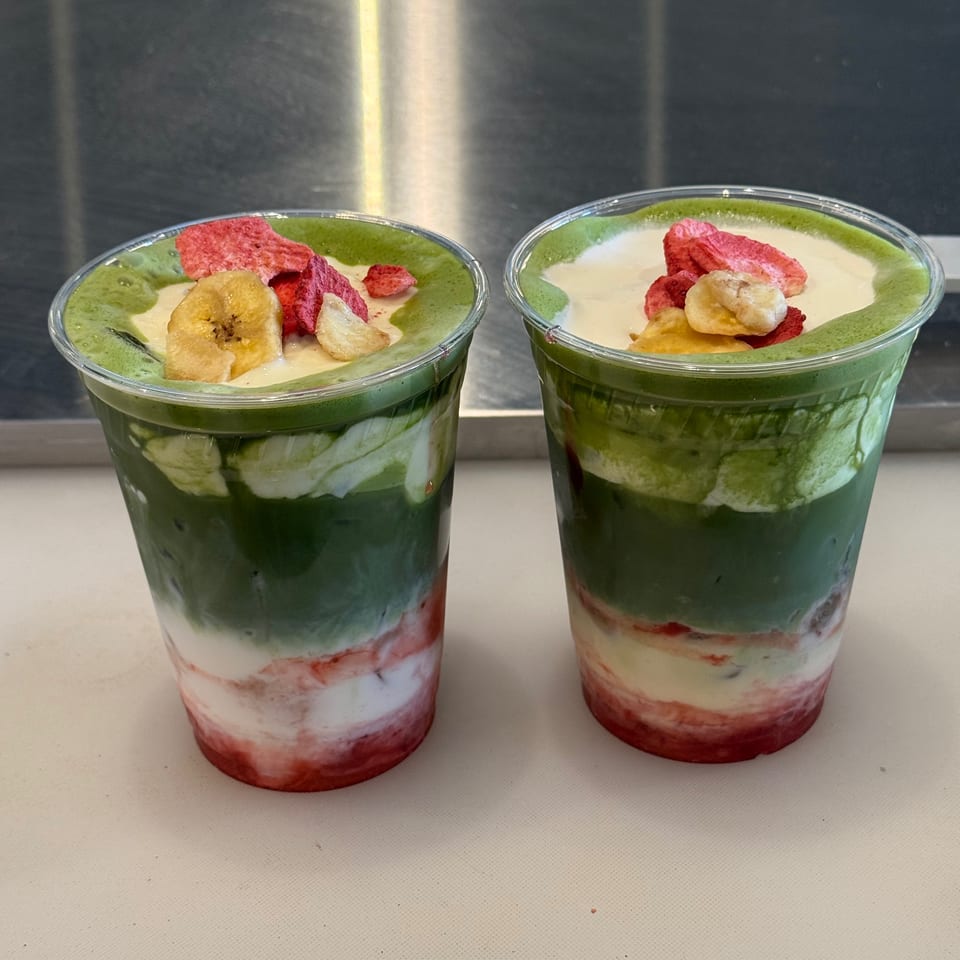 Two layered matcha drinks with cream, strawberry, and banana chips in clear cups.