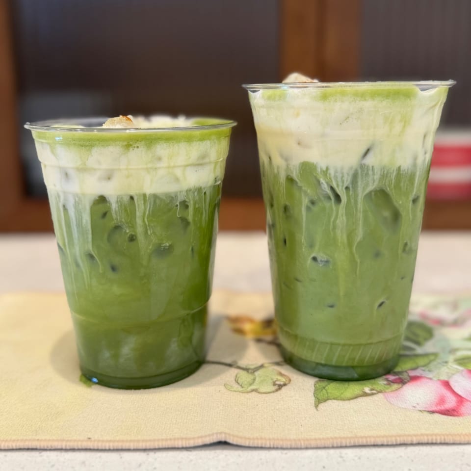 Two iced matcha lattes with cream tops and ice in clear plastic cups on a floral mat.