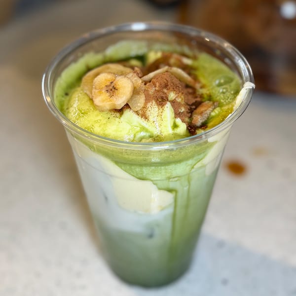 Iced matcha latte with whipped cream, banana slices, and cinnamon in a clear plastic cup.
