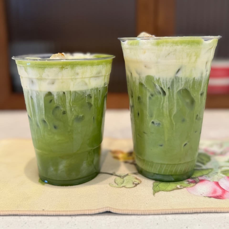 Two plastic cups of iced matcha lattes with creamy foam on a floral placemat.