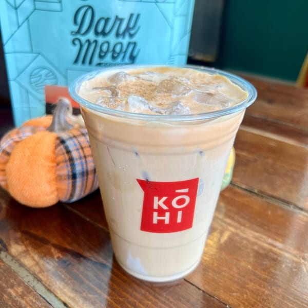 Iced latte with cinnamon in a KOHI Coffee cup, next to a plaid pumpkin decoration.