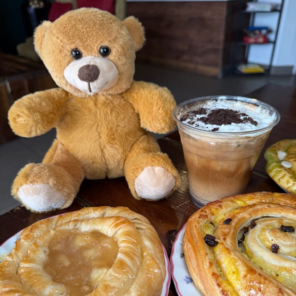 A teddy bear sits on a wooden table next to an iced coffee and two pastries.