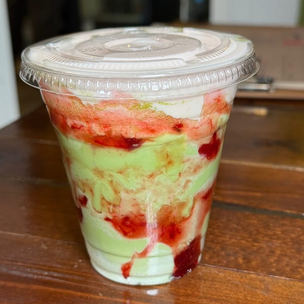 A vibrant green and red layered drink with whipped cream in a clear plastic cup on a wooden table.
