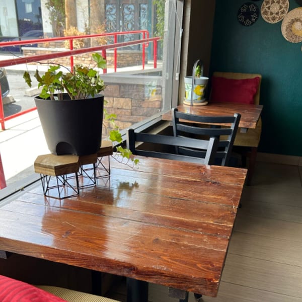 A cozy cafe interior with wooden tables, black chairs, and a potted plant by a large window.