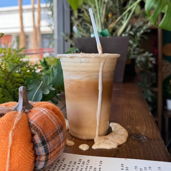 Iced coffee with foam spilling over, next to a plaid pumpkin and menu on a wooden table.
