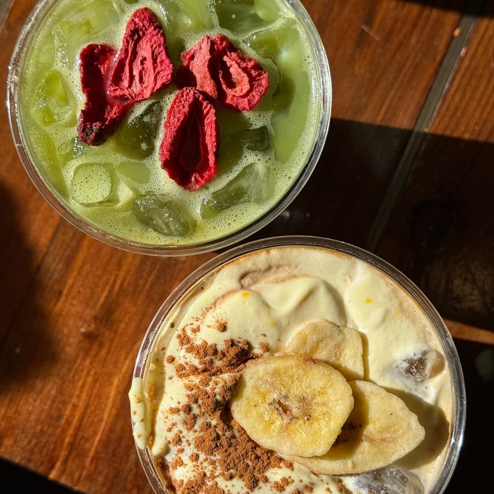 Two iced drinks on a wooden table: a green matcha with dried strawberries and a banana smoothie with cocoa.