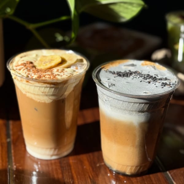 Two iced lattes, one topped with banana slices and cinnamon, the other with black sesame seeds.