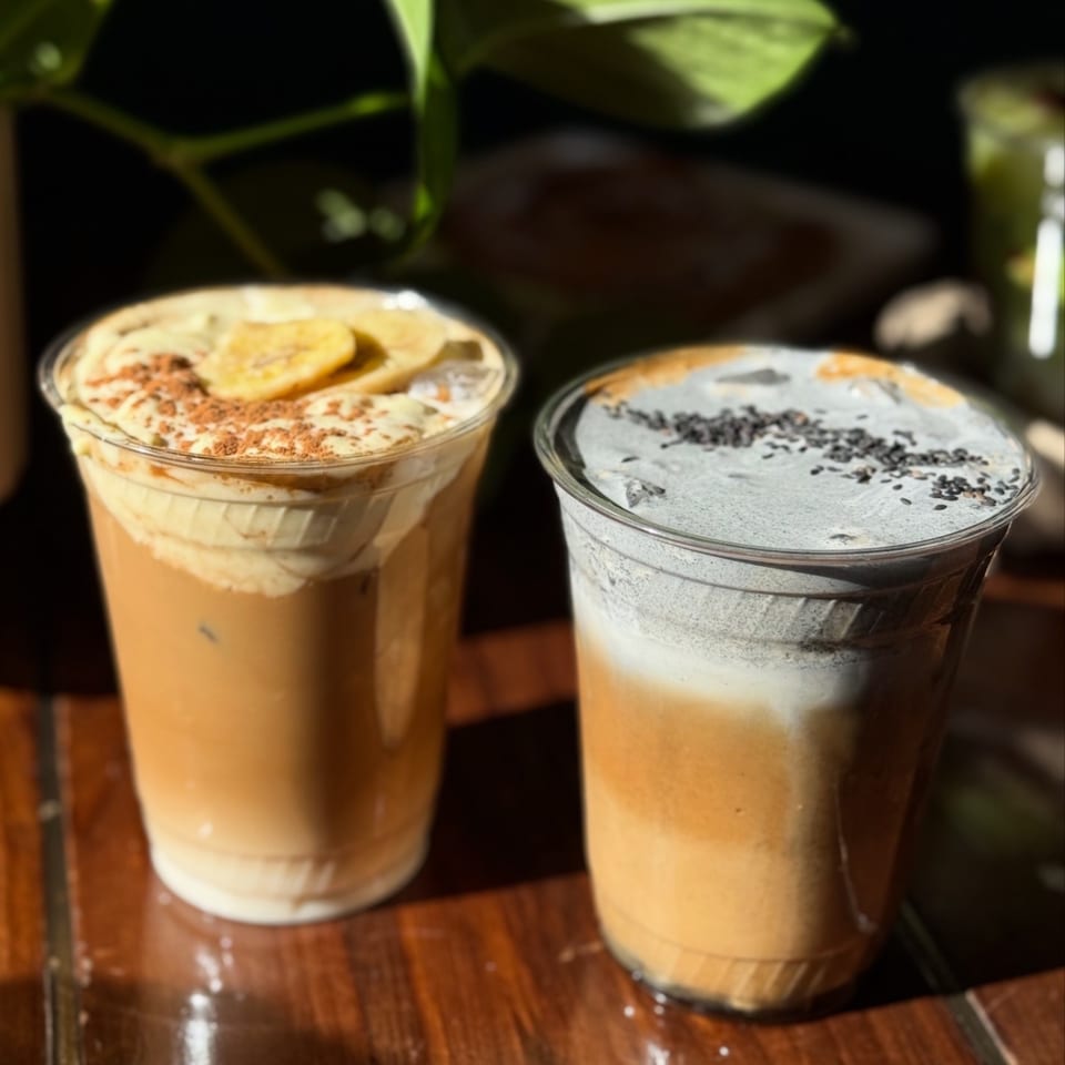 Two iced lattes, one topped with banana slices and cinnamon, the other with black sesame seeds.
