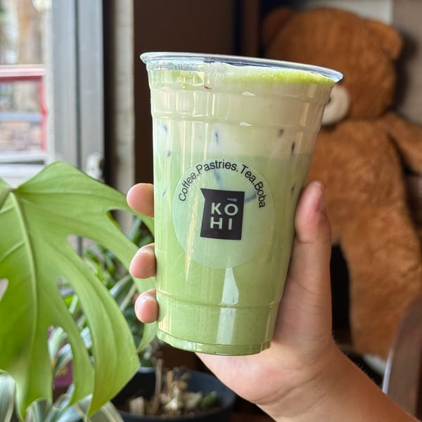Hand holding a KOHI Coffee iced matcha latte with a plant and teddy bear in the background.