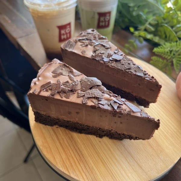 Two slices of KOHI Coffee chocolate cheesecake with chocolate shavings, and two iced drinks in the background.