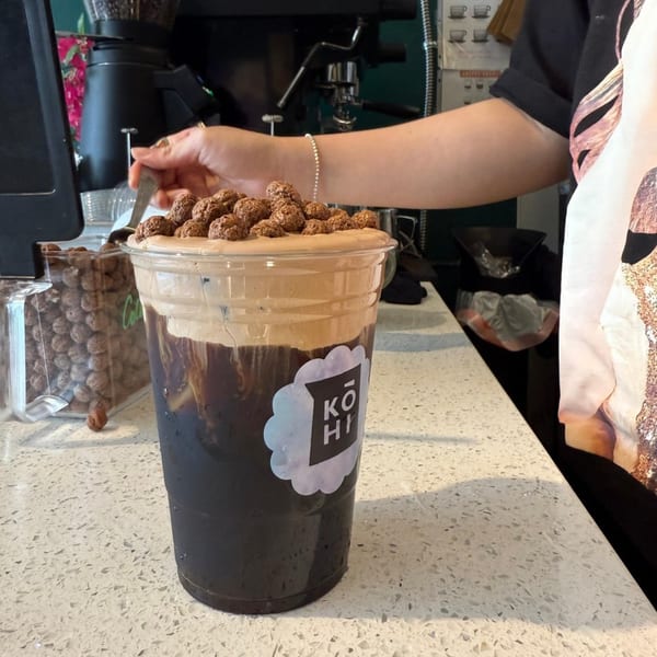 A KOHI Coffee drink with dark liquid, light foam, and chocolate cereal on top, being prepared by a barista.